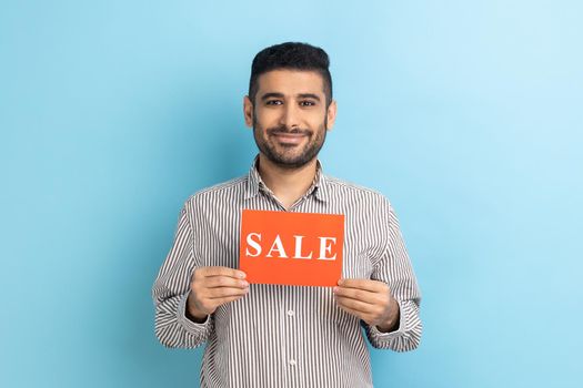 Smiling businessman holding card with Sale word, feeling carefree amused by discounts, Black Friday low prices, wearing striped shirt. Indoor studio shot isolated on blue background.