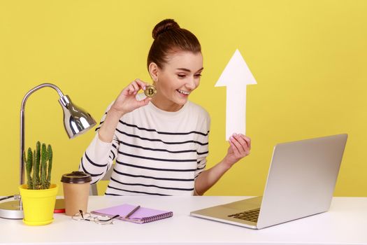 Positive woman showing golden bitcoin and white paper arrow to laptop screen, showing growth of crypto currency, sitting on workplace. Indoor studio studio shot isolated on yellow background.