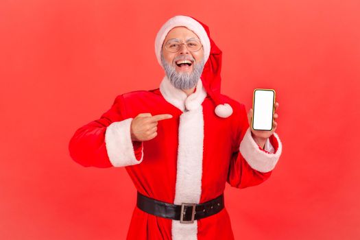 Portrait of excited elderly man with gray beard in santa claus costume standing with smart phone in hands, pointing at blank screen with index finger. Indoor studio shot isolated on red background.
