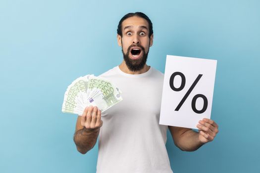 Portrait of astonished amazed man in white T-shirt holding paper with percent sign inscription and euro banknotes, looking at camera with open mouth. Indoor studio shot isolated on blue background.