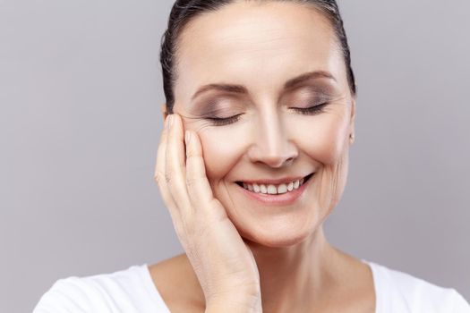 Closeup portrait of middle aged woman gently touching face with one hand and smiling, having healthy pure skin, female enjoying spa facial treatment. Indoor studio shot isolated on gray background.