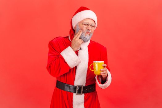 Portrait of elderly man with gray beard wearing santa claus costume suffering from terrible teeth pain after drinking hot or cold beverage. Indoor studio shot isolated on red background.