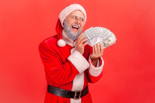 Portrait of elderly man with gray beard wearing santa claus costume with excited expression, greedy male with fan of dollars, looking at banknotes. Indoor studio shot isolated on red background.