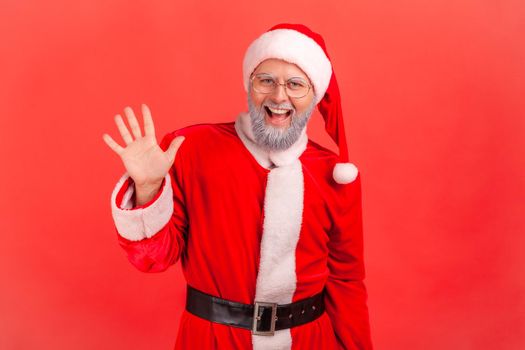 Hello. Happy elderly man with gray beard wearing santa claus costume raising palm to wave hi, greeting with hospitable friendly smile. Indoor studio shot isolated on red background.