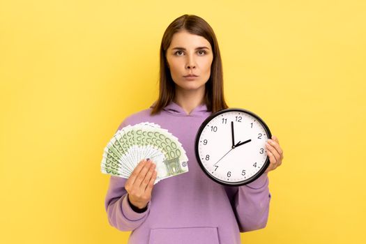 Portrait of rich dark haired woman holding big fan of euro banknotes and wall clock, time is money, looking seriously at camera, wearing purple hoodie. Indoor studio shot isolated on yellow background