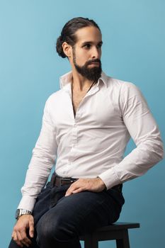 Portrait of attractive man with beard and dark collected hair wearing white shirt and black trousers, sitting and looking away with confident expression. Indoor studio shot isolated on blue background