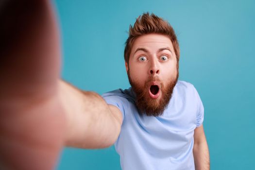 Portrait of shocked surprised handsome young adult bearded man standing with widely open mouth, making selfie POV, expressing astonishment. Indoor studio shot isolated on blue background.