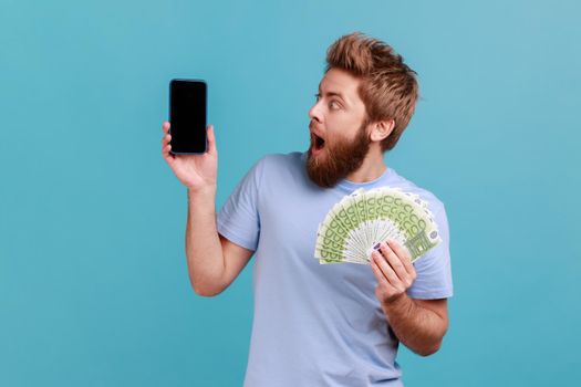 Portrait of astonished amazed bearded man holding euro banknotes and looking at cell phone with empty display, mock up for advertise. Indoor studio shot isolated on blue background.