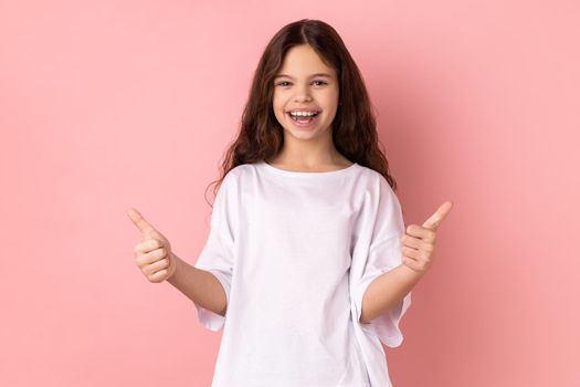 Portrait of smiling little girl wearing white T-shirt showing thumbs up, like gesture, looking at camera demonstrating her satisfaction. Indoor studio shot isolated on pink background.