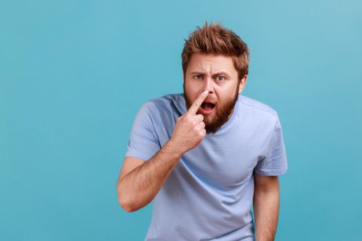 You are liar. Portrait of shocked bearded man standing, touching his nose and showing lie gesture, suspecting falsehood, dishonest talk. Indoor studio shot isolated on blue background.
