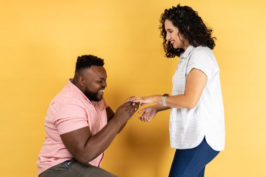 Portrait of happy lovely young couple in casual clothing standing together, romantic man puts on a wedding ring to her girlfriend, making proposal. Indoor studio shot isolated on yellow background.