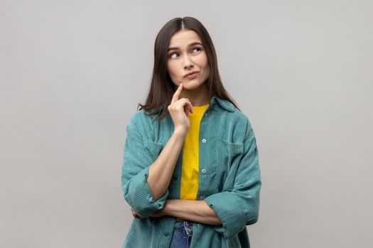 Portrait of pensive woman with dark hair thinking, looking away, having puzzled confused facial expression, wearing casual style jacket. Indoor studio shot isolated on gray background.