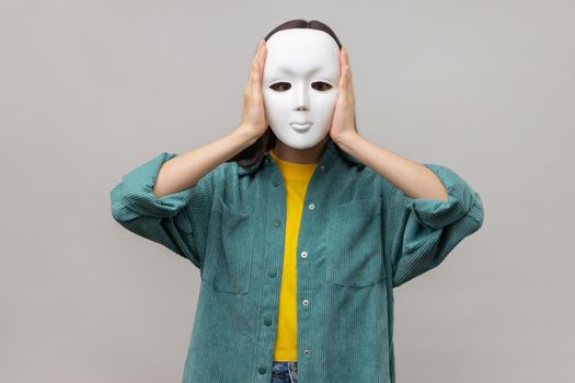 Portrait of unknown woman holding white mask, standing covering face, multiple personality disorder, wearing casual style jacket. Indoor studio shot isolated on gray background.
