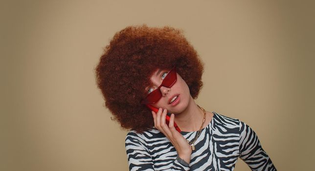 Tired young woman with brown lush wig making online conversation speaks on smartphone. Boring call. Blah expression. Nonsense talk. Adult stylish female girl isolated alone on beige studio background