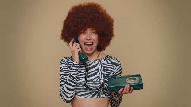 Hey you, call me back. Pretty young woman with brown lush wig talking on wired vintage telephone of 80s, says hey you call me back. Adult stylish female girl isolated on beige studio background indoor