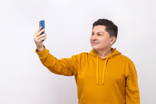 Portrait of cheerful man taking selfie using smartphone, talking on video call and smiling joyfully, mobile communication, wearing urban style hoodie. Indoor studio shot isolated on white background.