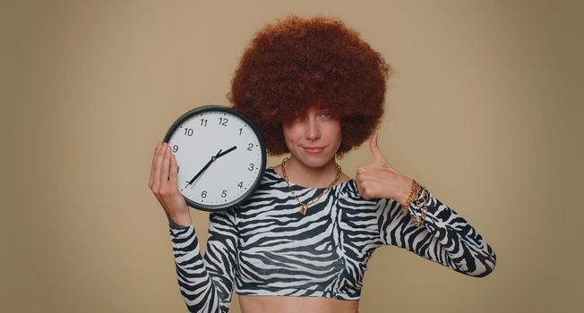 It is your time. Pretty young woman with brown lush wig showing time on wall office clock, ok, thumb up, approve, pointing finger at camera. Adult stylish female girl on beige studio background indoor