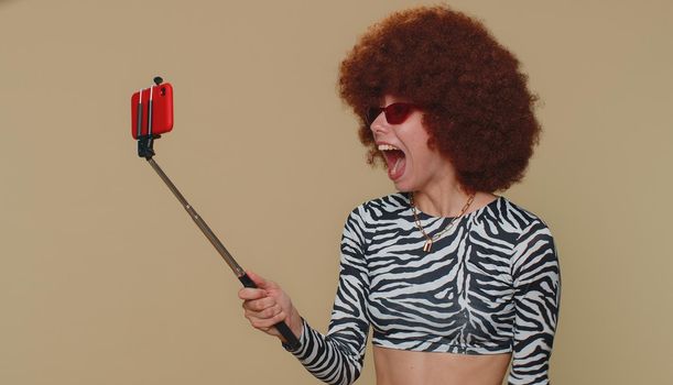 Wow reaction. Impressed pretty young woman with brown lush wig blogger taking selfie on mobile phone selfie stick, communicating video call online. Adult stylish girl on beige studio background indoor