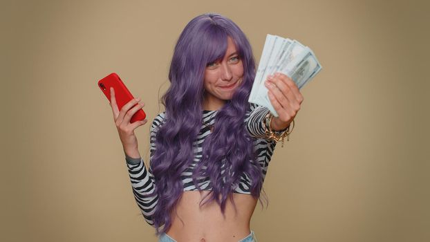 Happy young woman with lush wig looking smartphone display sincerely rejoicing win, receiving money dollar cash banknotes, success lottery luck. Adult stylish female girl on beige studio background