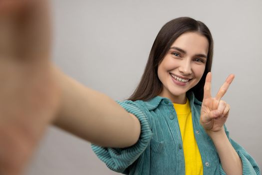 Smiling woman taking selfie POV, flirting, expressing positive emotions, showing v sign, victory gesture, wearing casual style jacket. Indoor studio shot isolated on gray background.