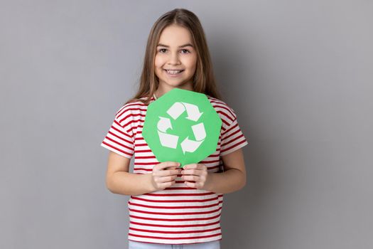 Portrait of charming responsible little girl wearing striped T-shirt holding in hands green recycling sing, ecology concept, looking at camera. Indoor studio shot isolated on gray background.