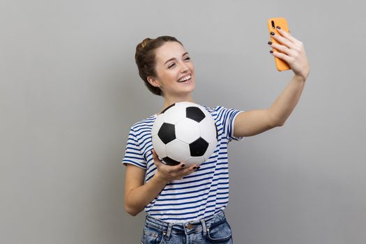 Portrait of smiling woman wearing striped T-shirt making selfie, having video call or broadcasting livestream with ball in hand. Indoor studio shot isolated on gray background.