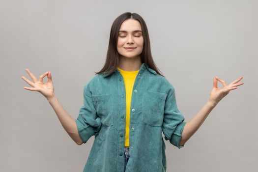 Portrait of peaceful young woman relaxing and doing meditation gesture with fingers, practicing yoga, wearing casual style jacket. Indoor studio shot isolated on gray background.