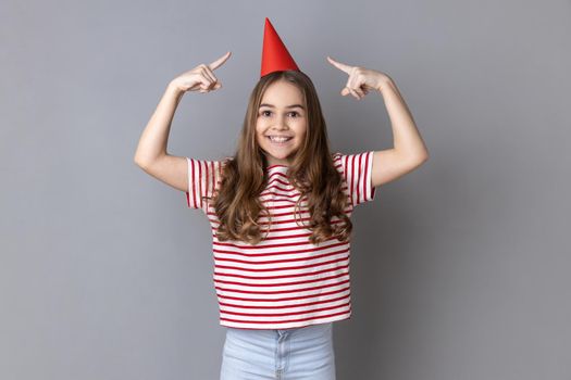 Portrait of little girl wearing striped T-shirt pointing at party cone on her head and looking at camera with toothy smile, celebrating birthday. Indoor studio shot isolated on gray background.