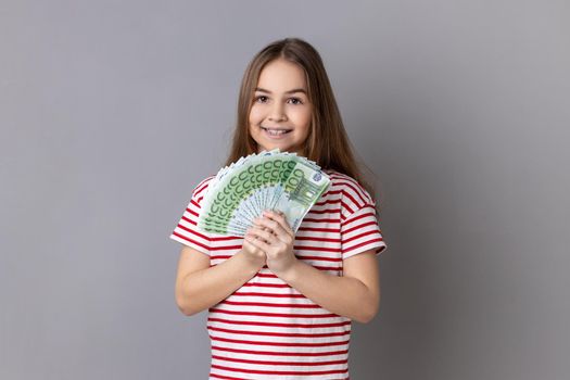 Portrait of satisfied little girl wearing striped T-shirt holding at fan of euro banknotes, showing big money and looking at camera. Indoor studio shot isolated on gray background.
