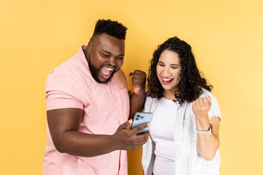 Portrait of excited extremely happy young couple in casual clothing standing together and looking at smart phone, clenched fists, celebrating victory. Indoor studio shot isolated on yellow background