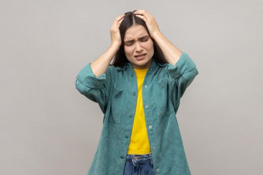 Portrait of unhealthy sick dark haired woman holding hands on head, feeling terrible headache, risk of migraine, wearing casual style jacket. Indoor studio shot isolated on gray background.