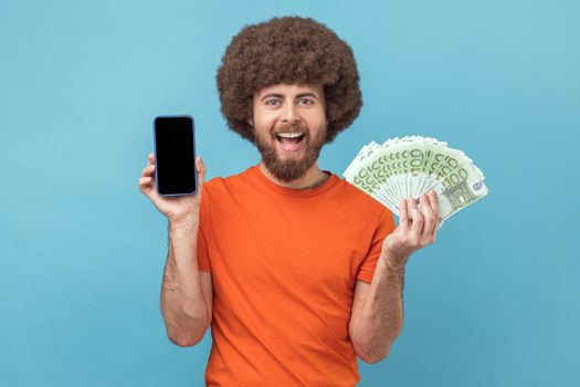 Portrait of satisfied man with Afro hairstyle wearing orange T-shirt showing euro banknotes and smart phone with empty display for promotional text. Indoor studio shot isolated on blue background.