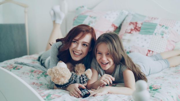 Laughing happy mother and cute daughter watching funny cartoon movie on TV while lying on bed at home in morning have fun at cozy bedroom