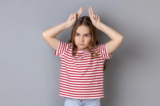 Portrait of aggressive bully little girl wearing striped T-shirt showing bull horns gesture over head, frowning as before attack. Indoor studio shot isolated on gray background.