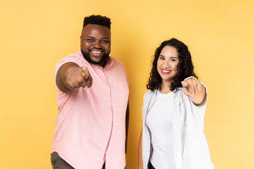 Portrait of smiling satisfied couple in casual clothing standing together and pointing at camera with index fingers, expressing positive emotions. Indoor studio shot isolated on yellow background.
