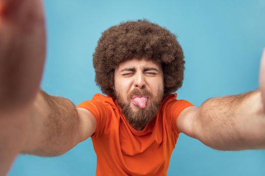 Portrait of cheerful childish man with Afro hairstyle wearing orange T-shirt taking selfie with closed eyes and sticking tongue out, point of view. Indoor studio shot isolated on blue background.