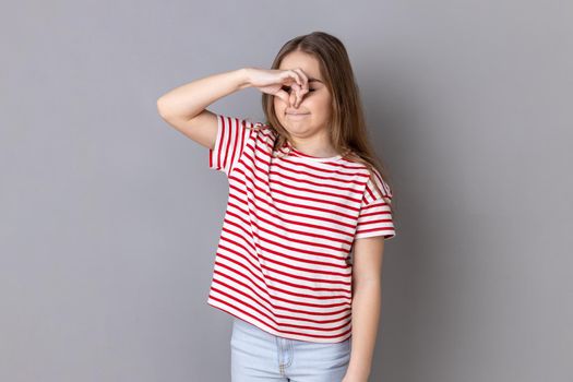 Portrait of confused little girl wearing striped T-shirt pinching her nose to hold breath, feeling repulsion and disgust to unpleasant odor. Indoor studio shot isolated on gray background.