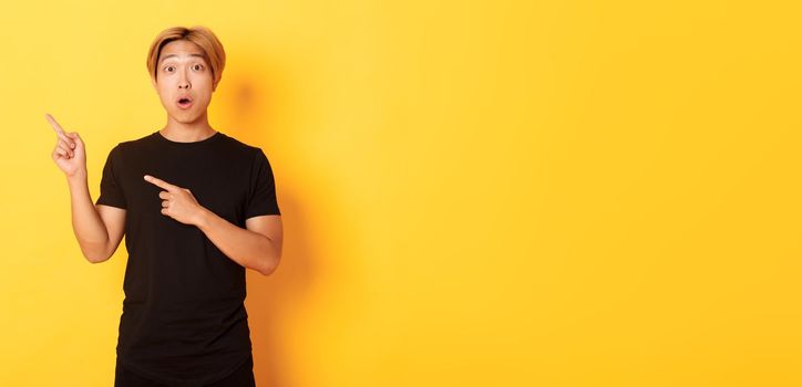 Portrait of impressed and excited handsome asian guy in black t-shirt, react to your logo, pointing fingers upper left corner.