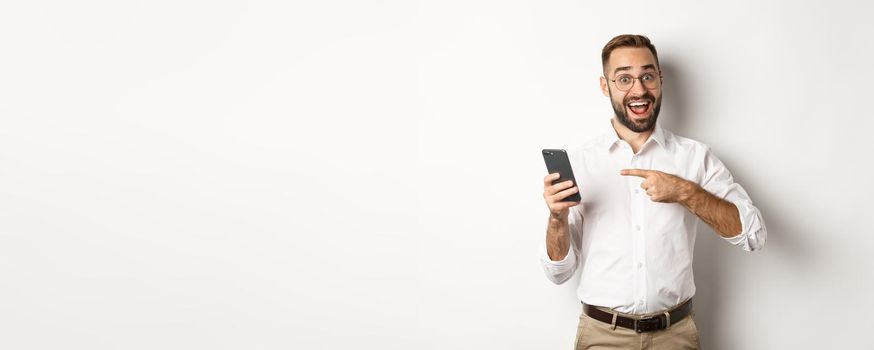 Man looking excited and pointing finger at mobile phone, showing good online offer, standing over white background.