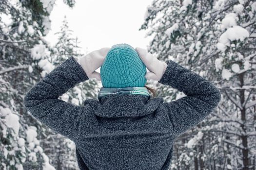 portrait of a girl in a gray coat and a blue knitted hat stands in a beautiful winter snowy forest on a cloudy snowy day, raising her hands to her head, examines the trees. Back view