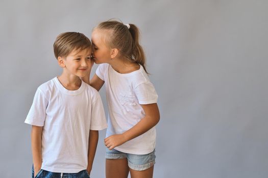 Cute two kids, little boy and girl in white t-shirts, best friends. Girl whispering secrets into boy's ear. on gray background with copy space. Blank white t-shirt for your design