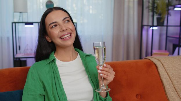Happy caucasian girl hold glass of champagne cheering drinking, celebrate success win, career grow up, lucky situation at home in living room. Young woman celebrating rent or purchase of new house