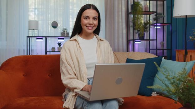 Lovely adult girl freelancer enters the home living room and sits down on orange couch, opens laptop. Young woman works on notebook, sends messages, makes online purchases, watching movies, working