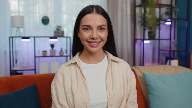 Close-up portrait of happy smiling caucasian girl in shirt, looking at camera, celebrate good news. Young woman indoor isolated at home in living room sitting on orange couch. Female nature beauty