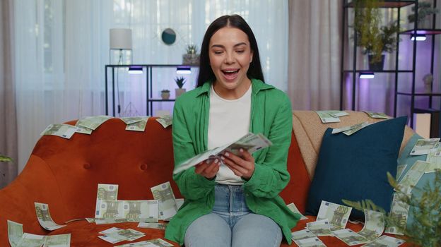 Successful happy rich business woman girl counting money cheering up with high profits, lottery game win, satisfied of wealth income earnings salary, on home room couch full of euro cash banknotes