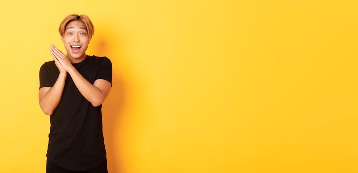 Portrait of happy and surprised asian man clap hands and smiling delighted, standing yellow background.