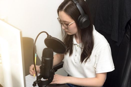 Young woman with glasses and headphones taking notes and preparing for podcast recording in front of microphone, professional record audio, technology and media concept