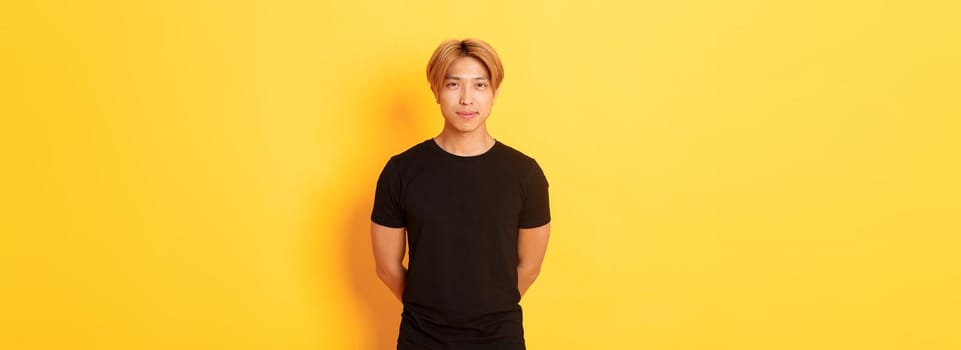 Portrait of handsome smiling asian blond guy looking sassy, holding hands behind back, standing yellow background.