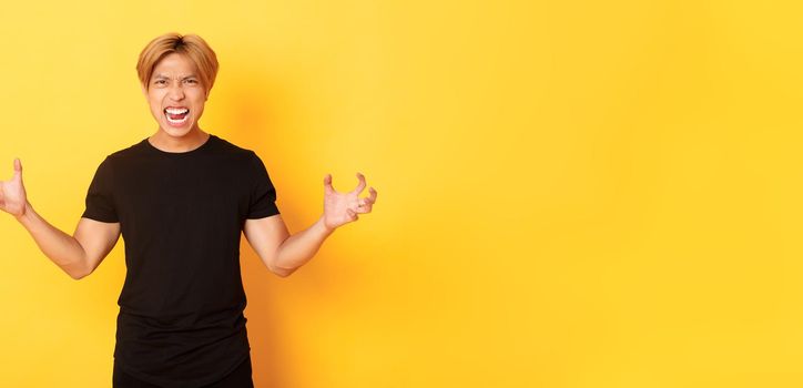 Portrait of outraged, pissed-off asian man shaking hands hateful and grimacing, shouting angry, standing over yellow background.