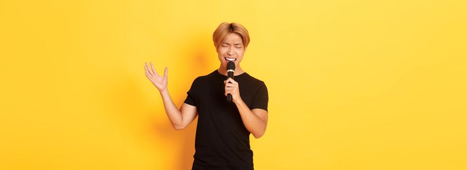 Handsome asian male singer, korean guy singing song at karaoke in microphone with passion, standing over yellow background.